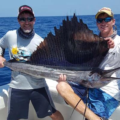 Two men on a boat holding a large sailfish above the water.