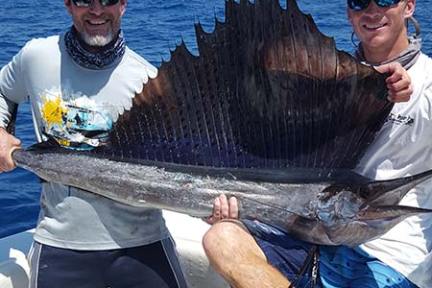 Two men on a boat holding a large sailfish above the water.