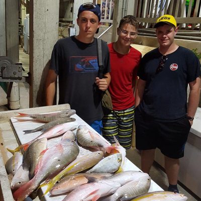 Three people standing by a table with assorted fish on it, under a wooden structure at night.