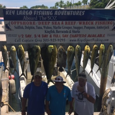Three people stand under a fishing sign with caught fish hanging above them, near a boat dock.