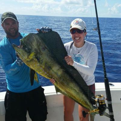Two people on a boat holding a large mahi-mahi fish, with ocean in the background.
