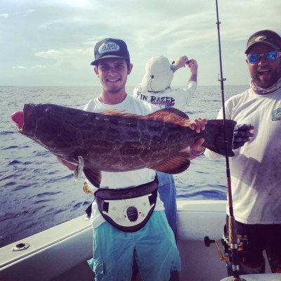 Three people on a boat; one holding a large fish horizontally, others smiling, ocean in the background.
