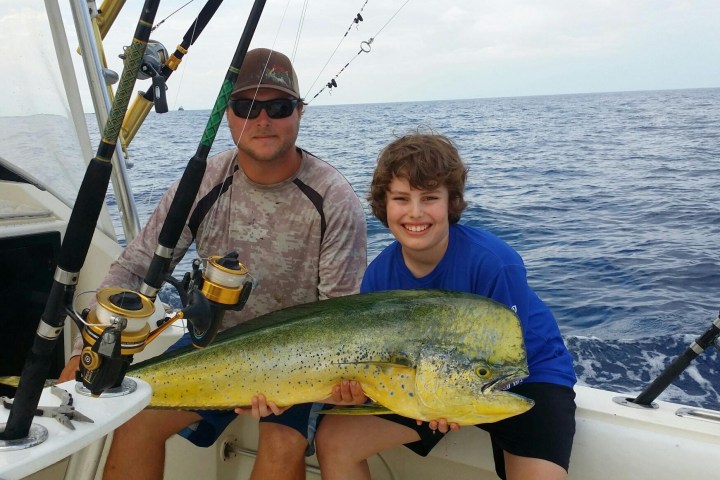 Two people on a boat holding a large fish, with fishing rods around and the ocean in the background.