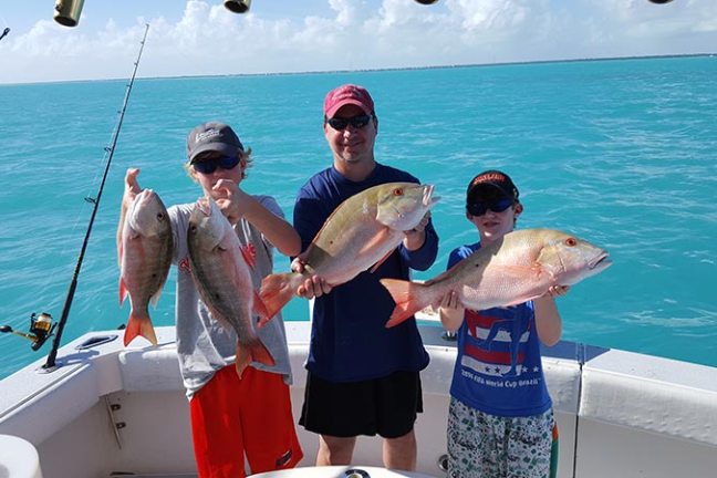 Three people on a boat holding large fish over turquoise water, showcasing a successful fishing trip.
