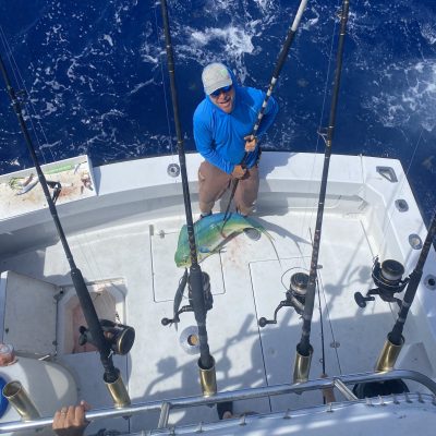 Man on boat holding fishing rod with fish on deck, surrounded by ocean.