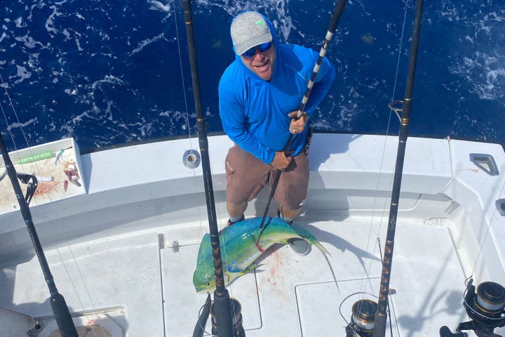 Person on a boat holding a fishing rod with a large fish on deck, ocean in the background.