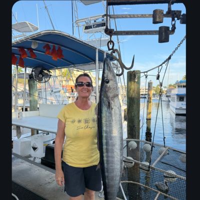 Person in yellow shirt standing next to a large fish hanging vertically near boats and water.