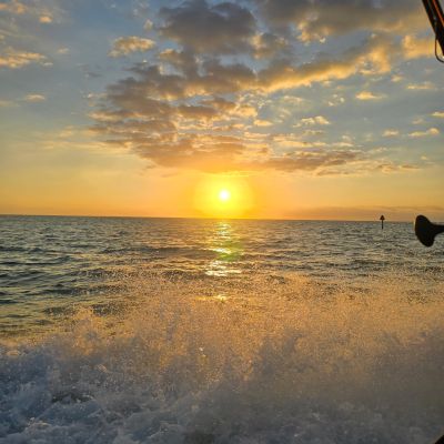 Sunset over ocean with waves splashing, cloudy sky, and sunlight reflection on water.
