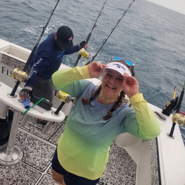 Two people on a boat with fishing rods, one smiling, wearing a colorful shirt and cap.