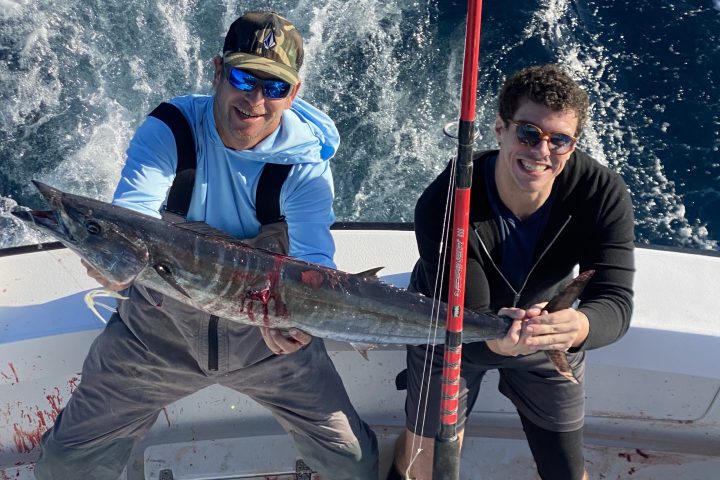 Two people on a boat holding a large fish with fishing pole in foreground.