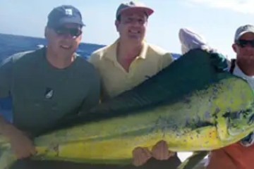 Three people on a boat holding a large fish against a backdrop of ocean and sky.