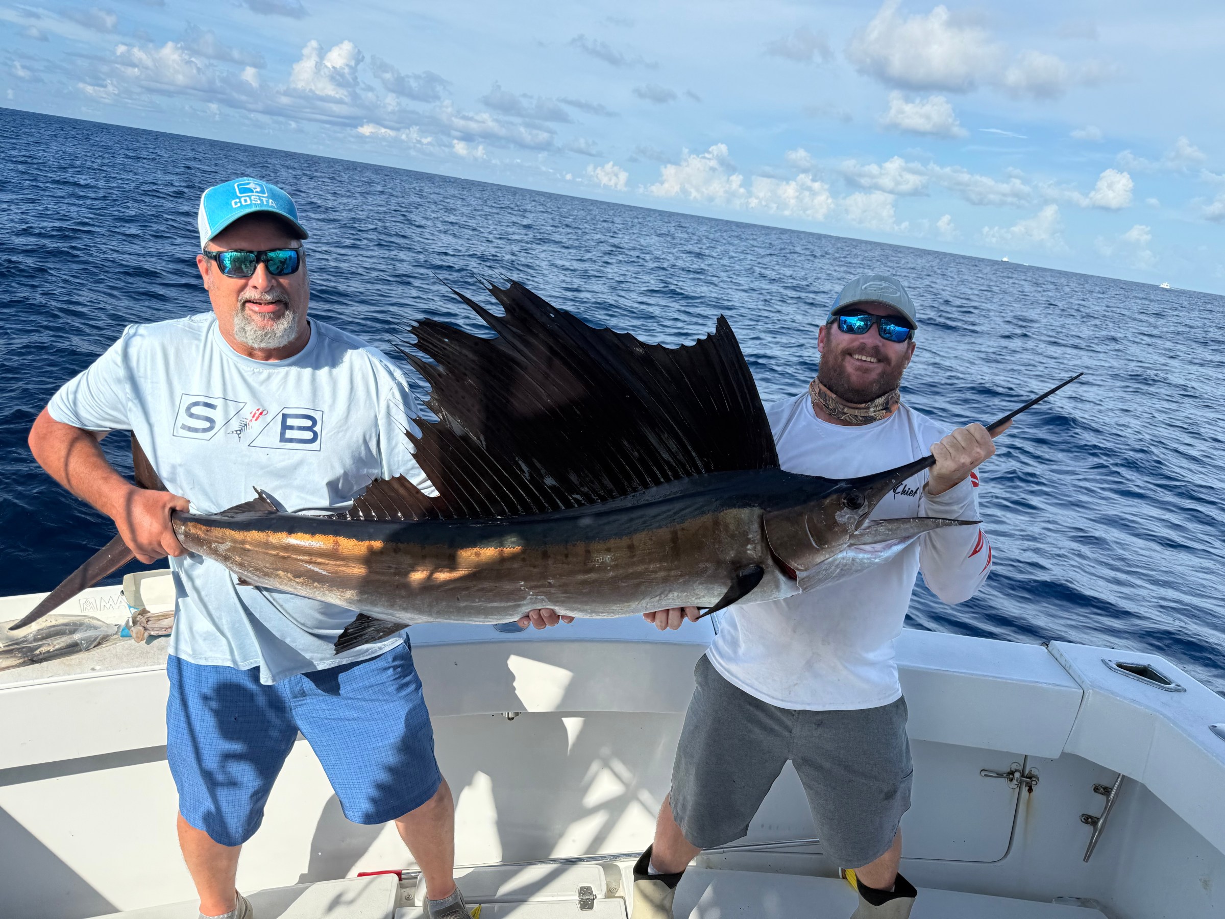 Two men on a boat holding a large sailfish, with the ocean in the background.