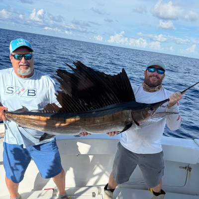 Two men on a boat holding a large sailfish, with the ocean in the background.