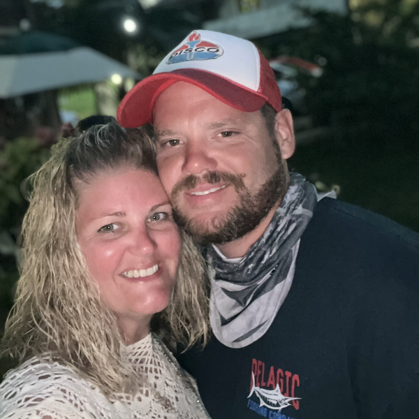 A smiling couple poses outdoors in casual clothing at dusk.