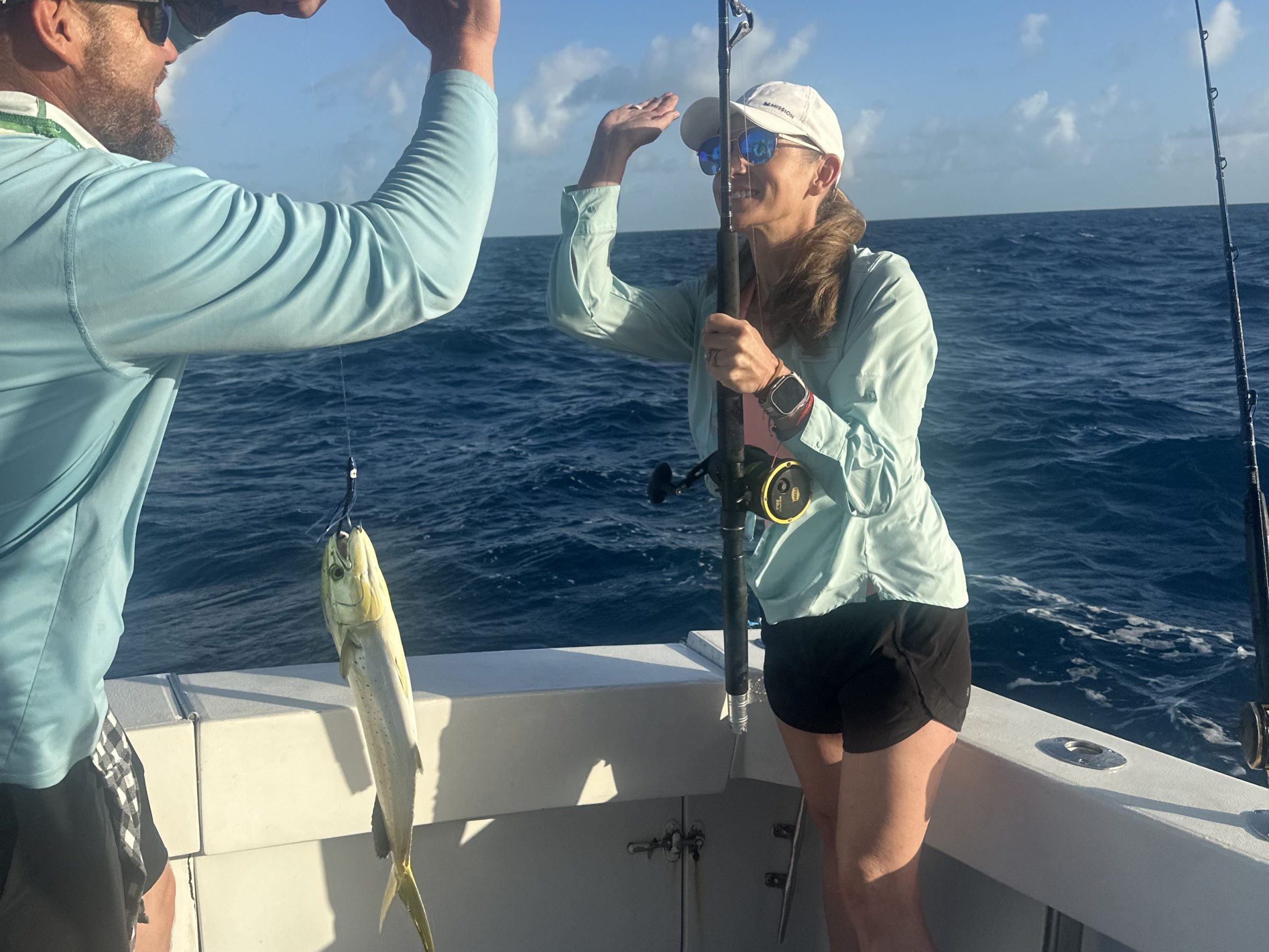 Two people on a boat high-fiving near a caught fish, with ocean in the background.