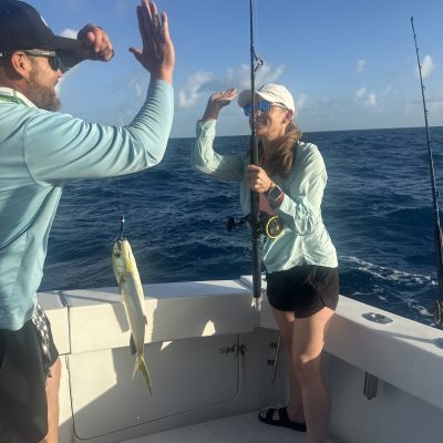 Two people on a boat high-fiving near a caught fish, with ocean in the background.