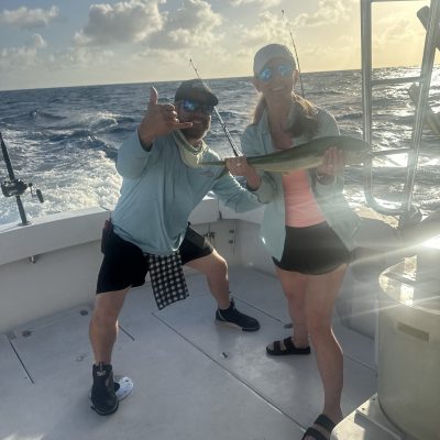 Two people on a boat, one holding a fish, with ocean and sunset in the background.
