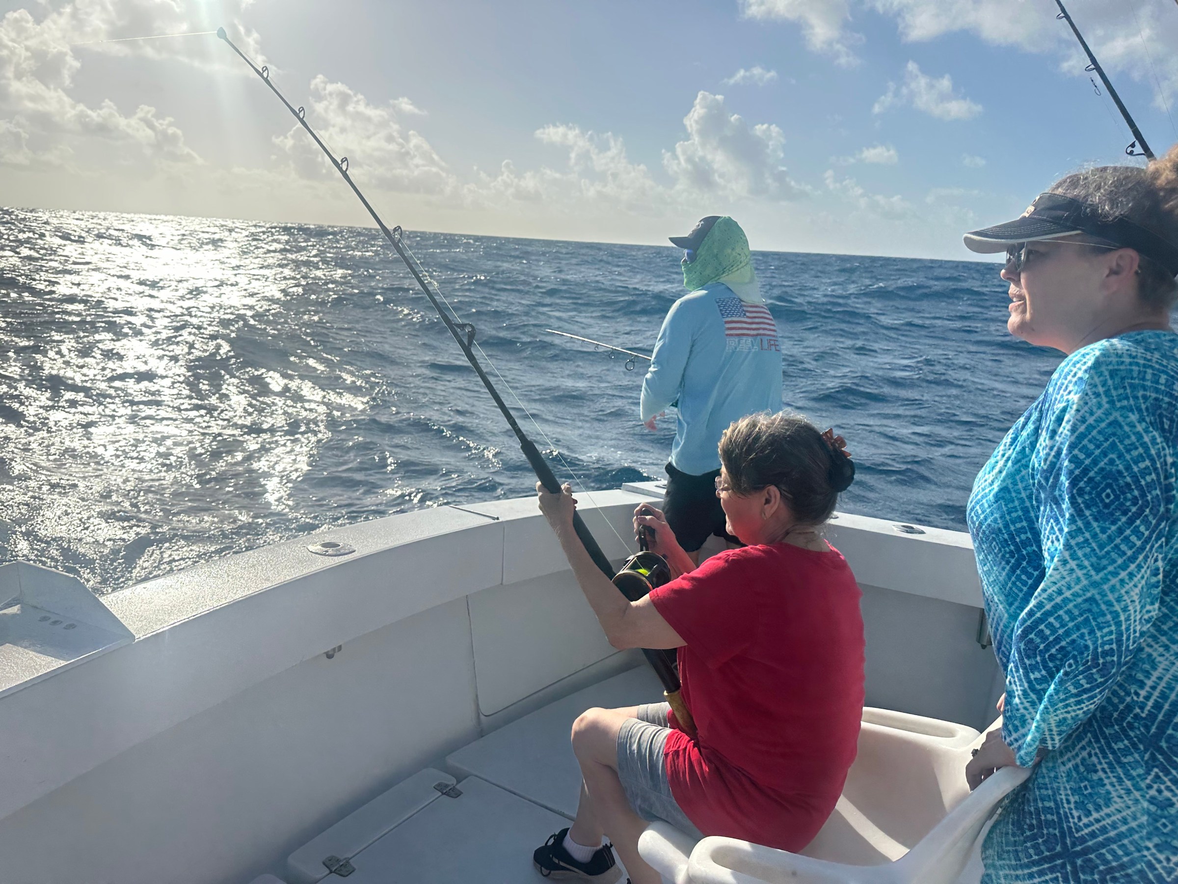 Three people fishing on a boat under a sunny sky with ocean waves visible.