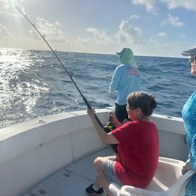 Three people fishing on a boat under a sunny sky with ocean waves visible.
