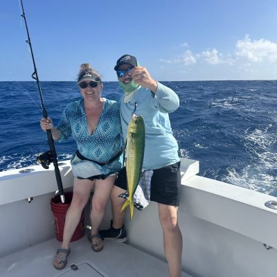 Two people on a boat holding a fishing rod and a caught fish with the ocean in the background.