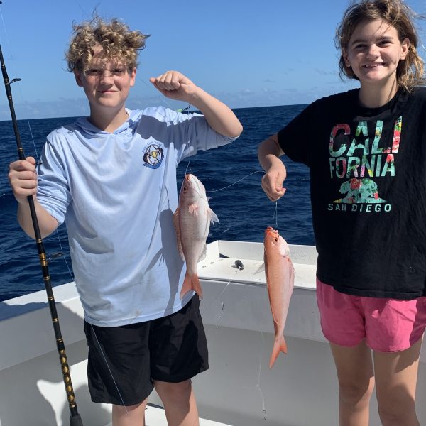 Two kids on a boat holding fishing rods and caught fish, with the ocean in the background.