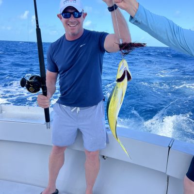 Person on a boat holding a fishing rod and a caught fish, smiling with a blue ocean backdrop.
