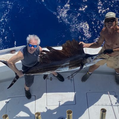 Two people on a boat holding a large sailfish with bright blue ocean in the background.