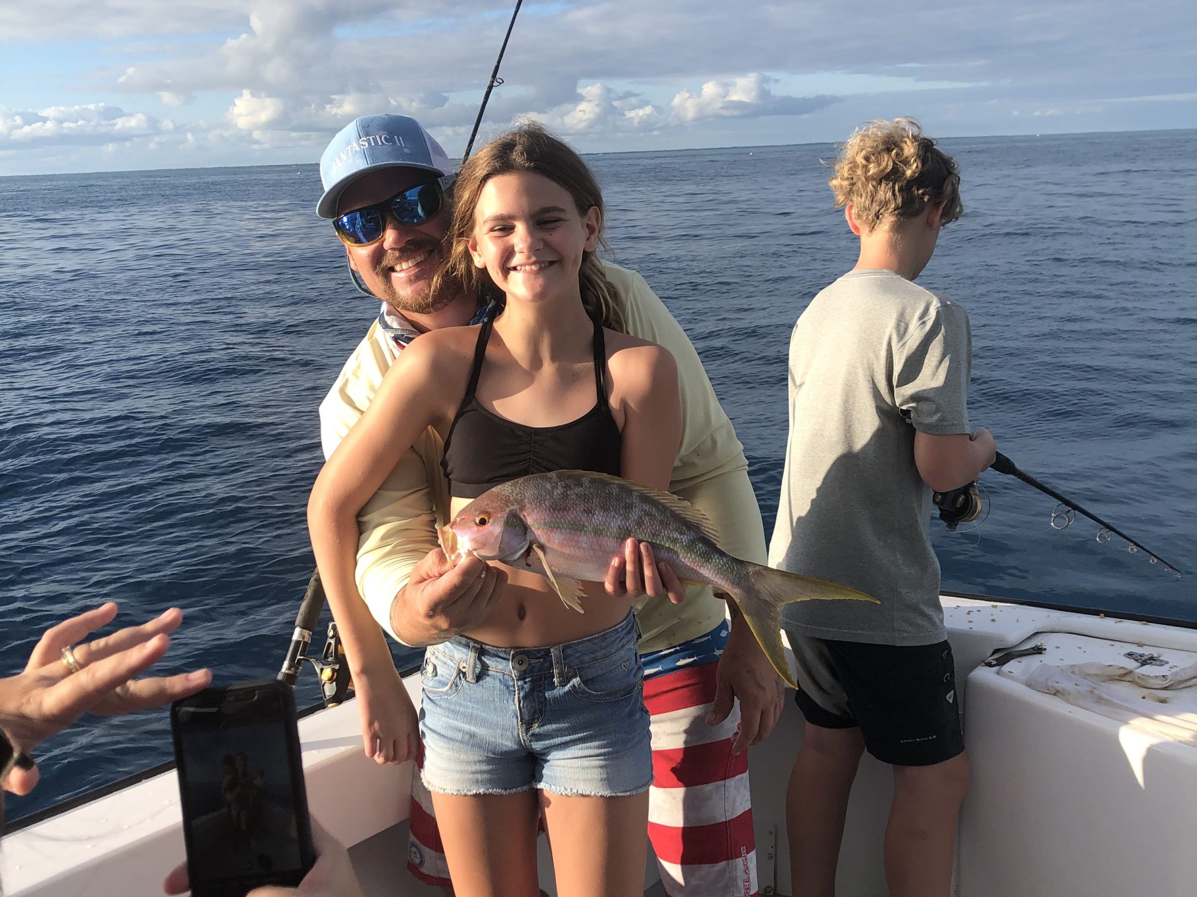 Girl holds fish with adult on boat, another child fishes, phone captures moment.