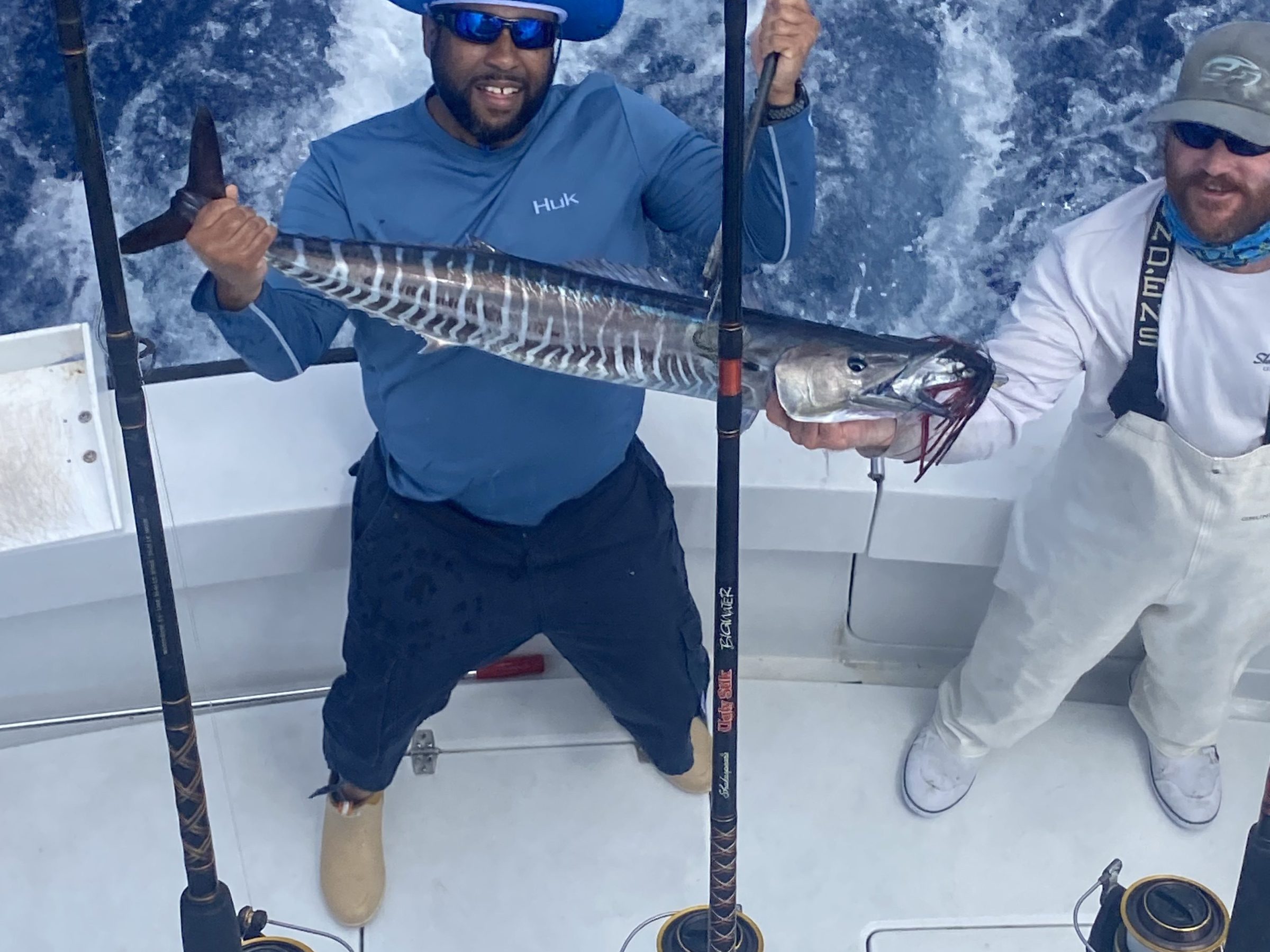 Two men on a boat holding a large fish, with fishing rods and ocean waves in the background.