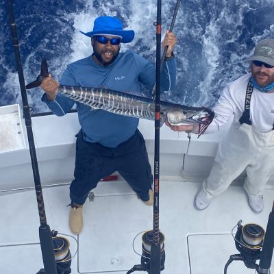 Two men on a boat holding a large fish, with fishing rods and ocean waves in the background.