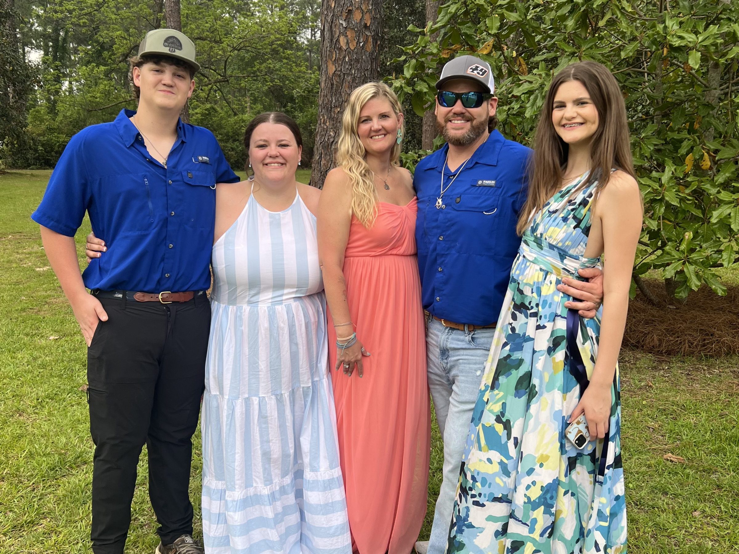 Five people standing outdoors, smiling, in colorful dresses and shirts, surrounded by trees.
