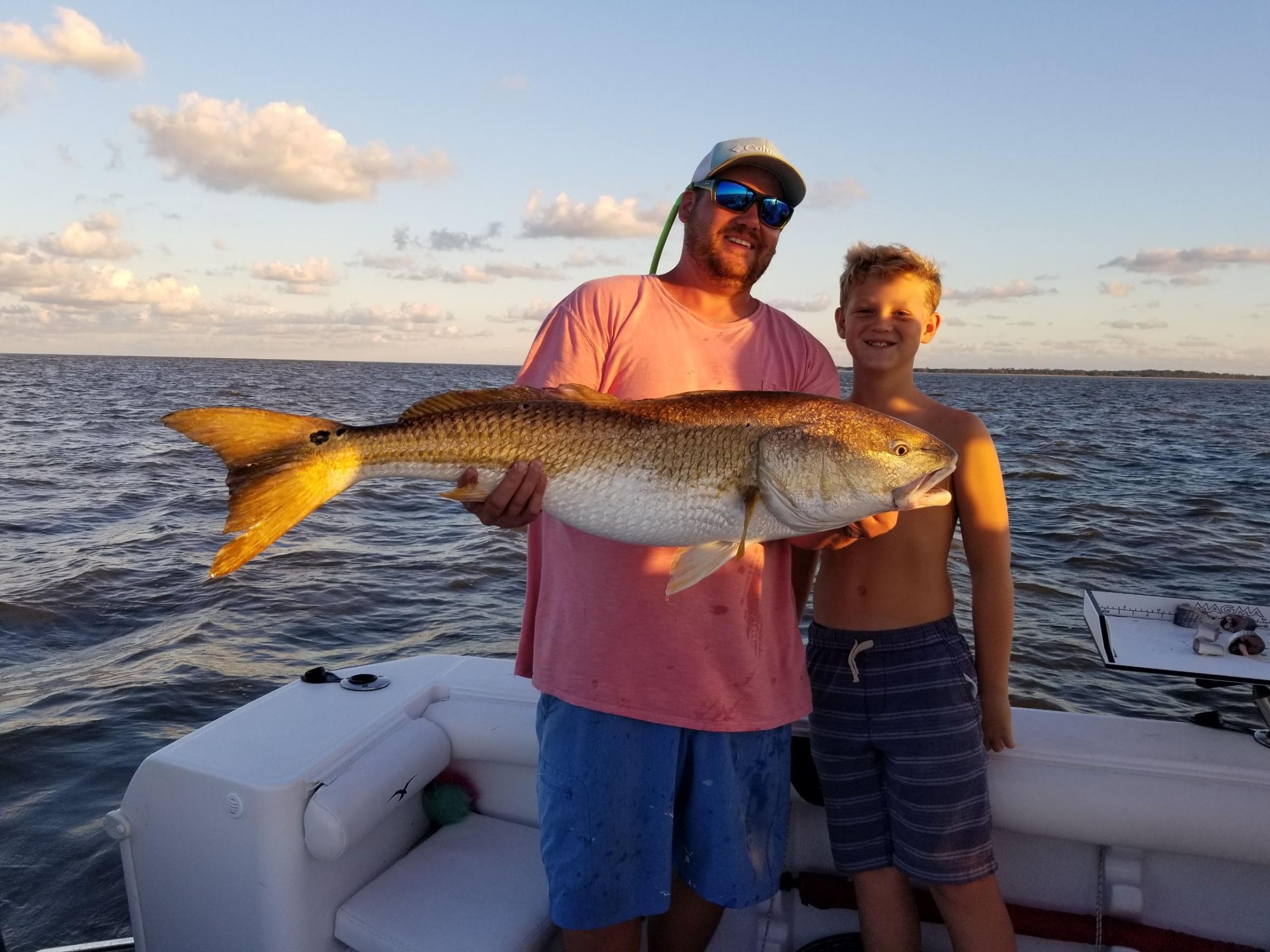 Two people on a boat holding a large fish with water and sky in the background.