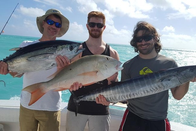 Three men on a boat holding large fish in sunny weather.