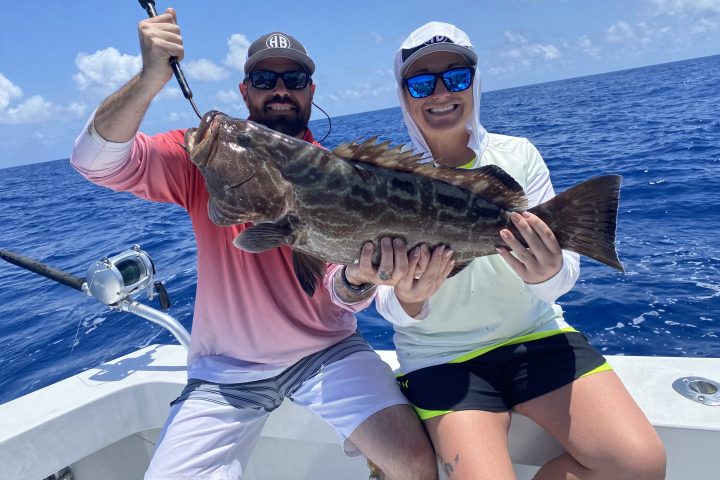Two people on a boat holding a large fish with blue ocean and sky in the background.
